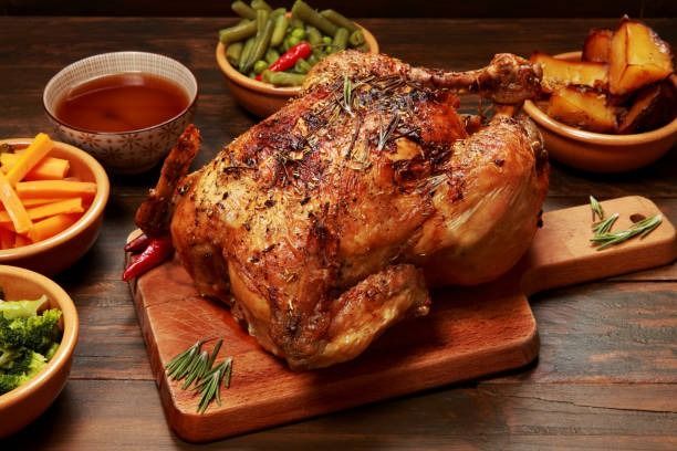 Pollo entero asado sobre una tabla de madera con guarnición de verduras y salsa en una mesa rústica.