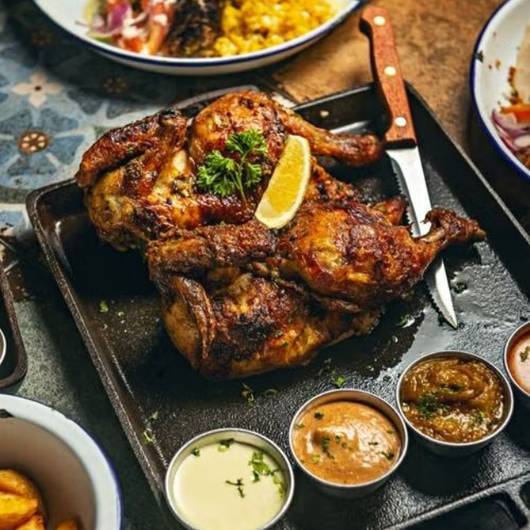 Pollo a la parrilla en una fuente con rodaja de limón, cuchillo y tres salsas para mojar en una mesa.
