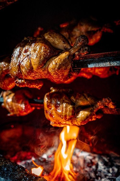 Pollo asándose en un asador sobre llamas abiertas, con la piel carbonizada y el fuego incandescente debajo.