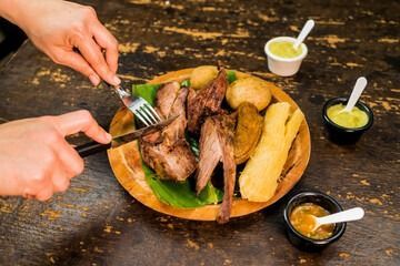 Manos cortando carne y patatas a la parrilla en una bandeja de madera, con pequeñas salsas para mojar cerca.