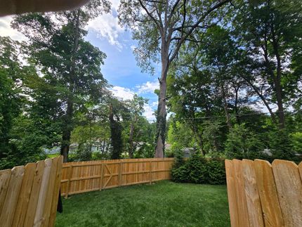 A view of a grassy backyard enclosed by a new wooden privacy fence, surrounded by tall, leafy green trees under a blue sky.