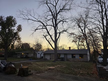 A single-story house with a yard at dusk, featuring several large, bare trees under a fading sunset sky.