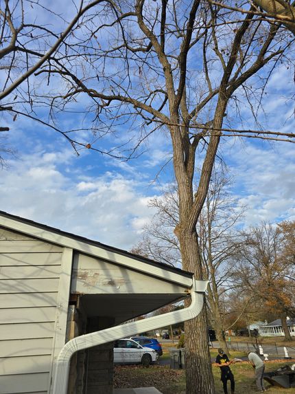 A house corner with a white gutter, a tall leafless tree nearby, and workers in the background under a cloudy blue sky.