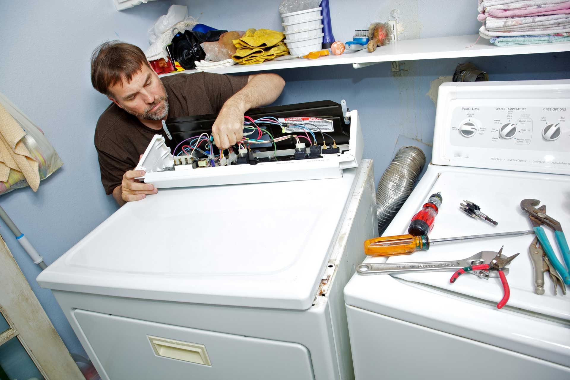 Man repairing a white dryer with tools laid out; cluttered laundry room.
