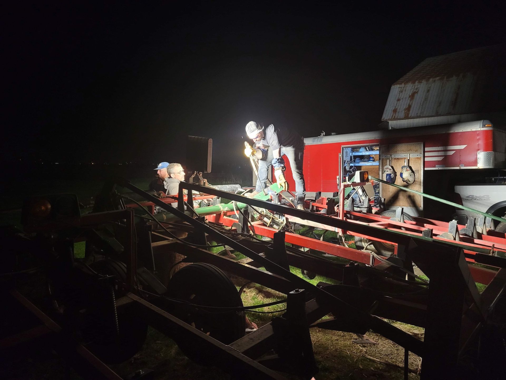 Des ouvriers réparent de nuit un tracteur agricole rouge, éclairé par une lumière vive.