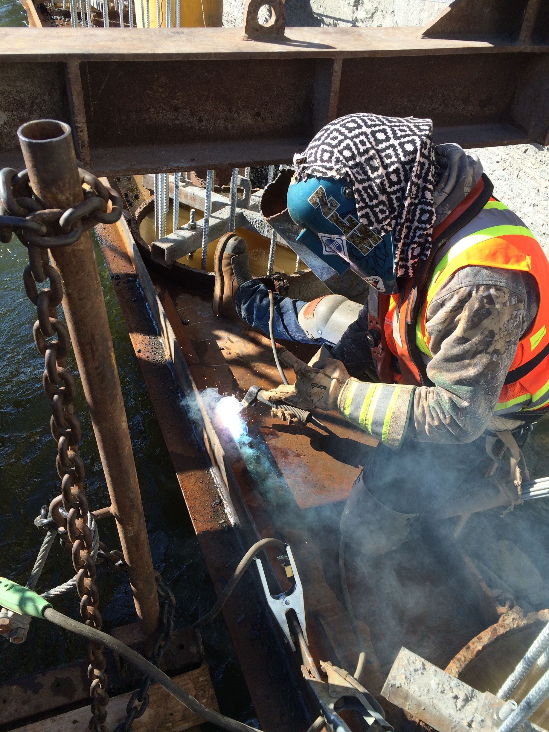 Soudeur en tenue de protection travaillant sur une structure métallique en extérieur, gilet de sécurité orange, étincelles visibles.