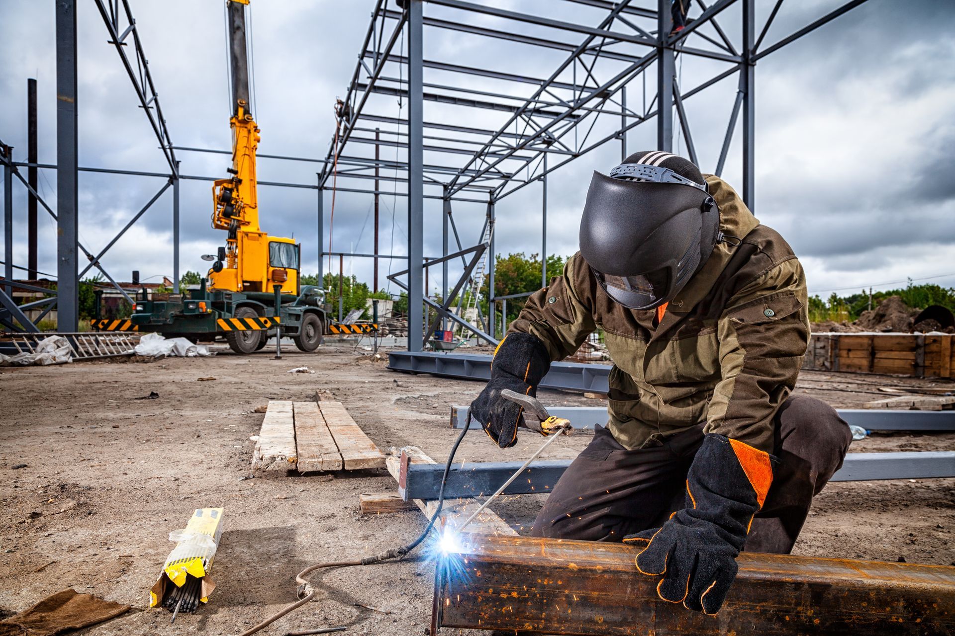 Un soudeur portant un casque soude des poutres métalliques sur un chantier de construction ; une grue est visible en arrière-plan.