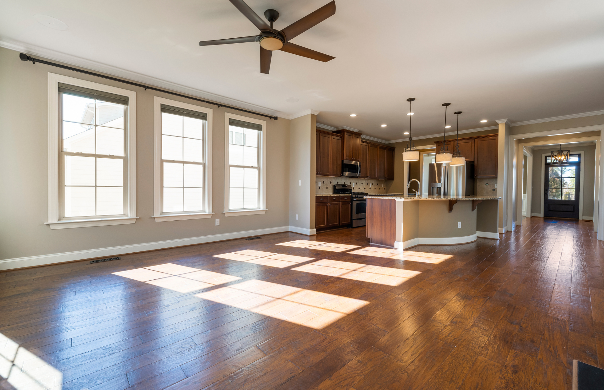 Empty open-plan living room and kitchen with hardwood floors, large windows, and a ceiling fan.