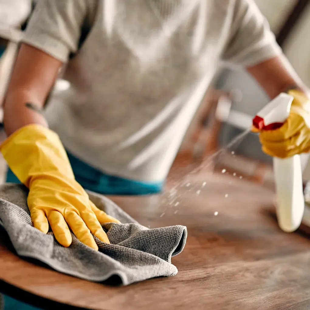 Person cleaning a wooden table with a gray cloth and spray bottle, wearing yellow gloves.