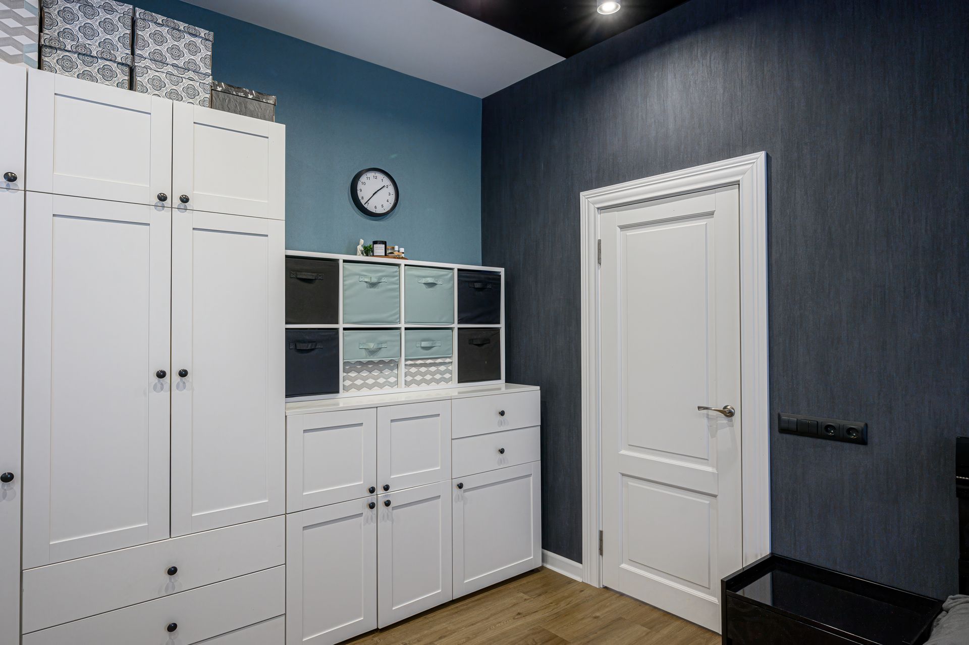 Small blue room with white storage cabinets, a wall clock, and a closed white door.