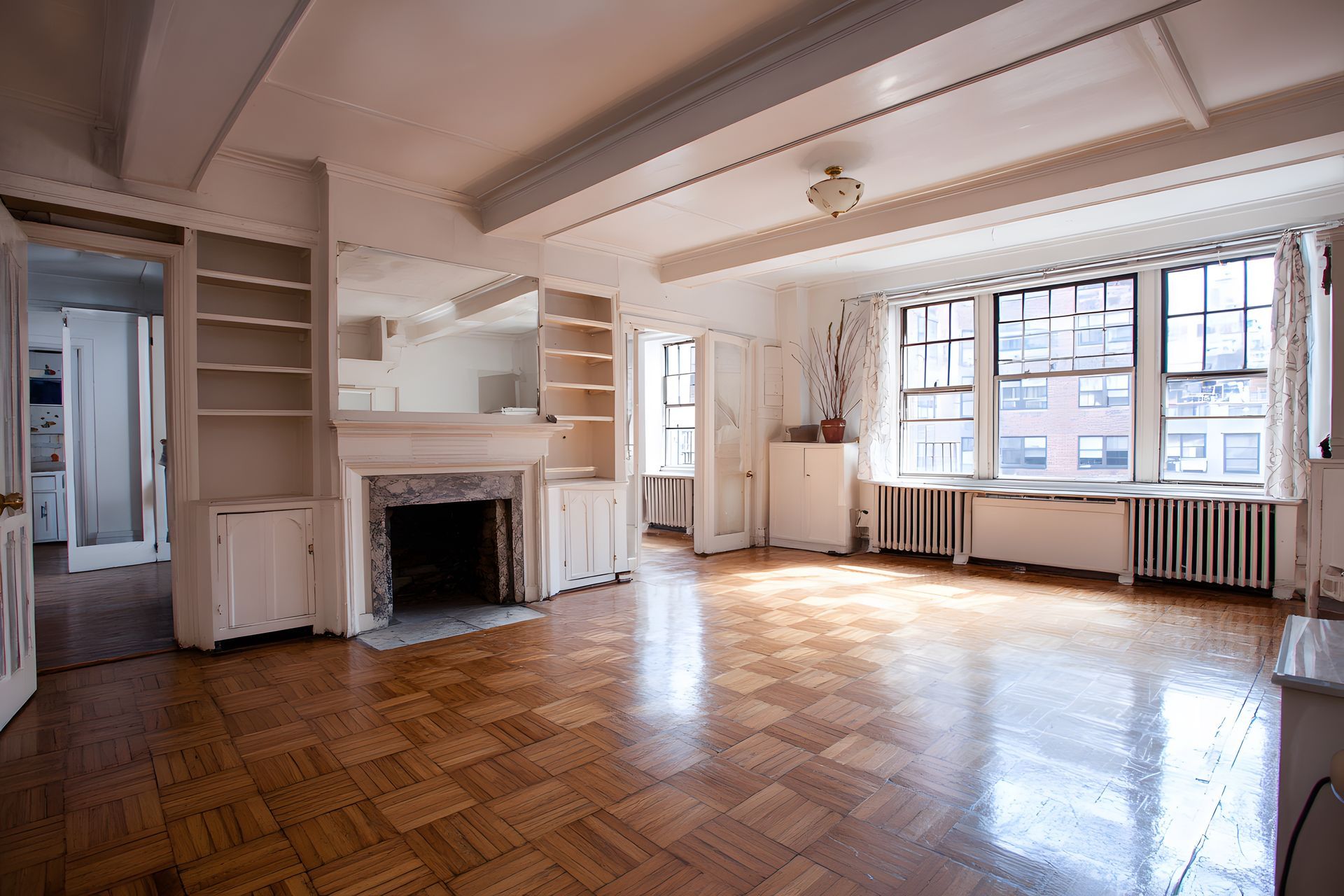 Empty sunlit living room with hardwood floors, fireplace, large windows, and white walls