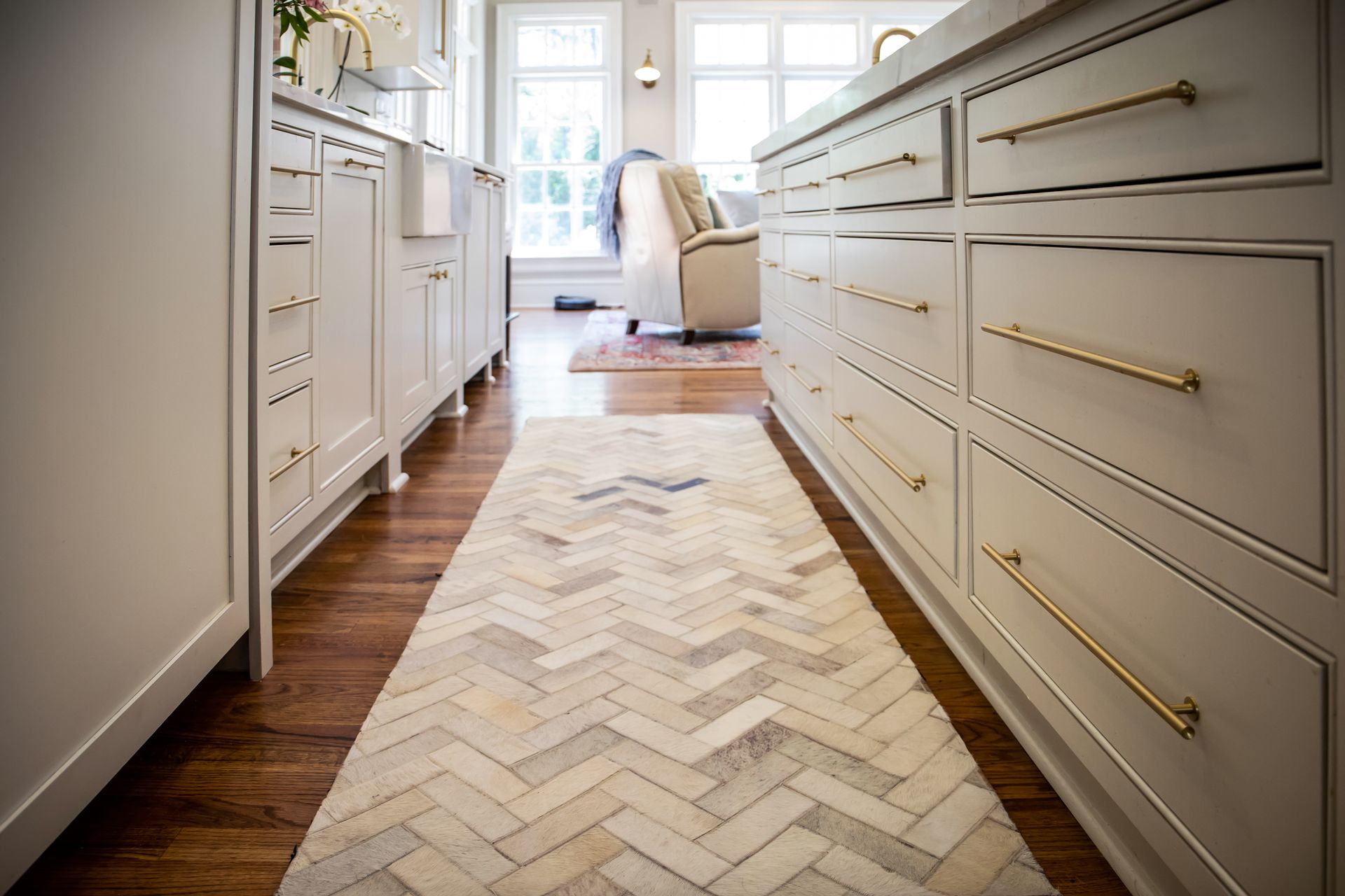 Narrow kitchen aisle with white cabinets and a herringbone wood floor leading to a bright living area