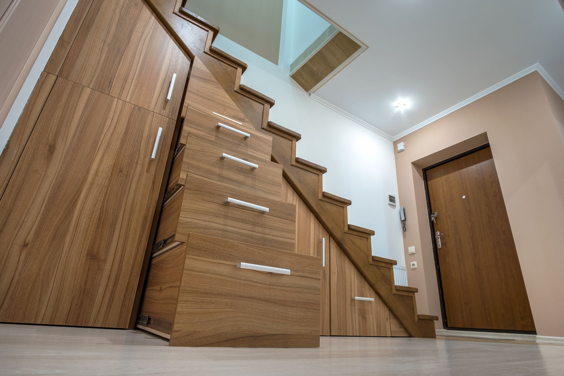 Wooden staircase with built-in drawers in a modern hallway, beside a closed door and a mirrored wall.