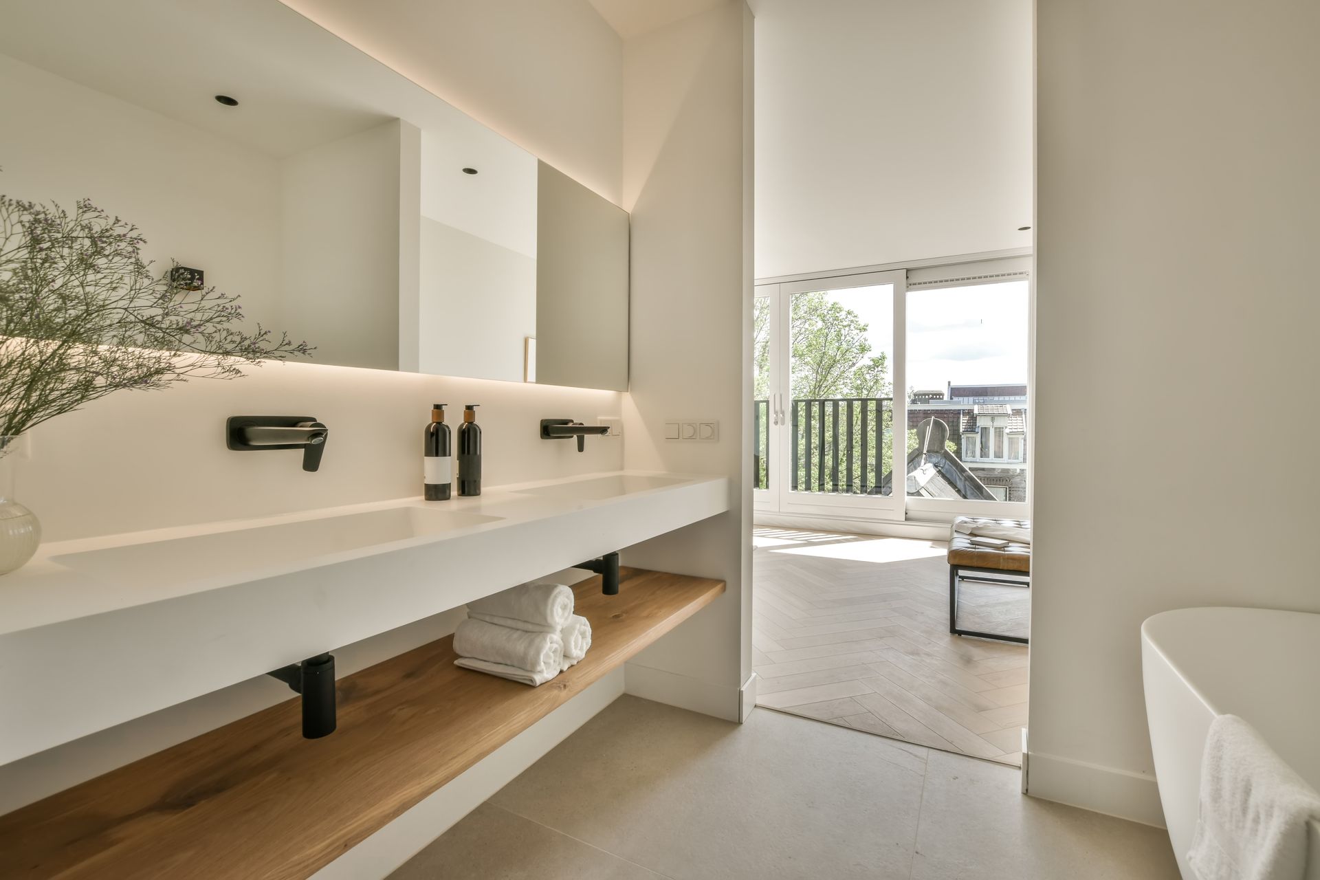 Modern bathroom with floating double sink, beige walls, and open balcony view beyond a bedroom doorway Modern bathroom with floating double sink, beige walls, and open balcony view beyond a bedroom doorway