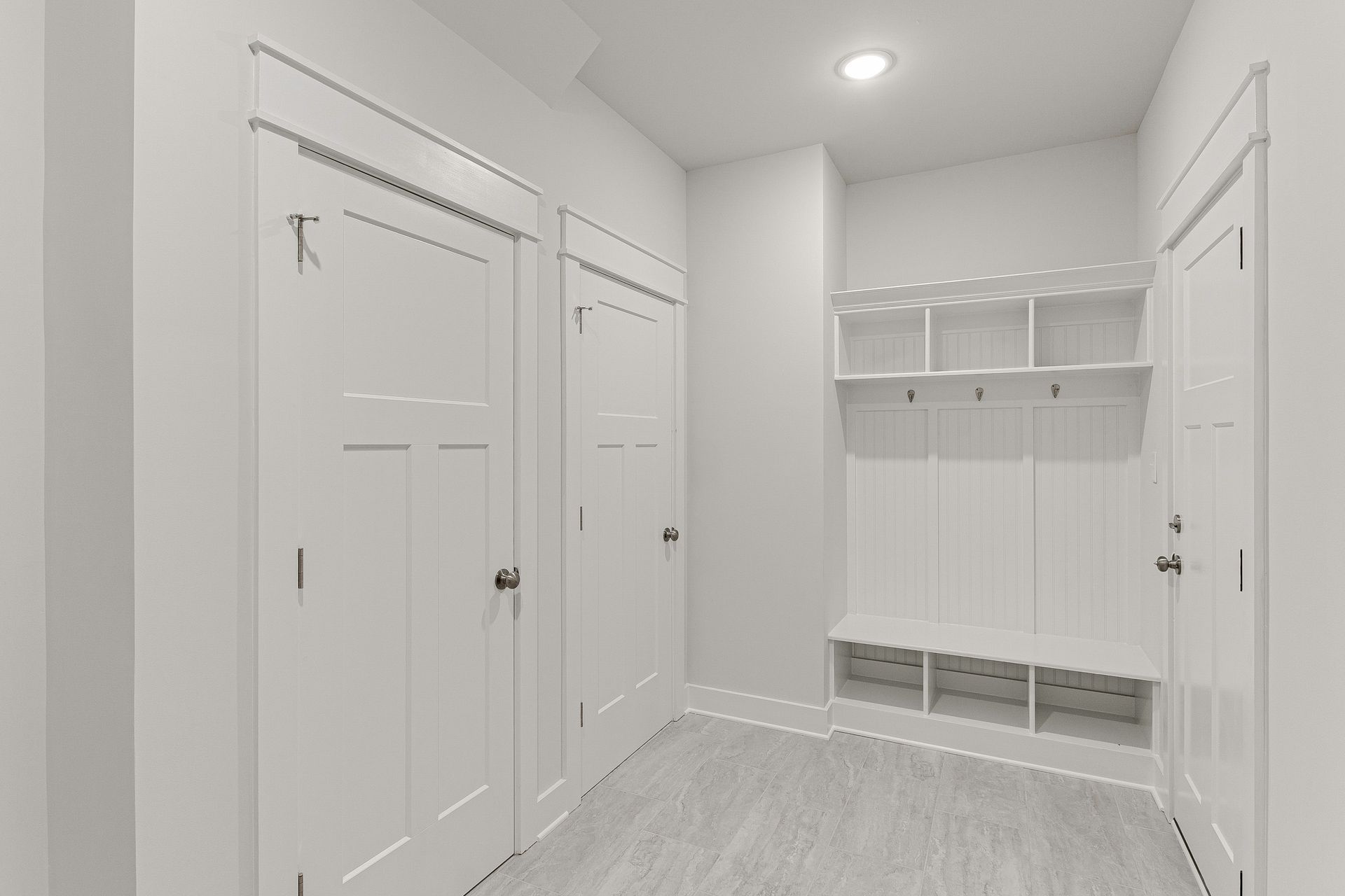 Bright white mudroom with hooks, shelves, cabinets, and two doors on a light wood floor.