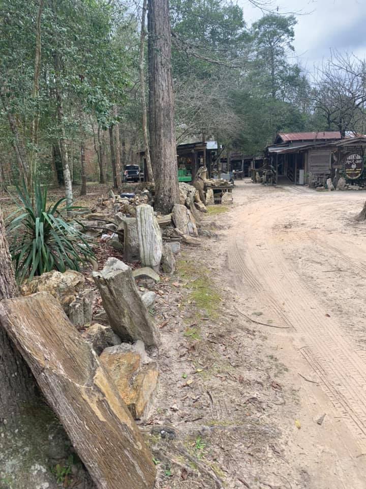 Dirt road beside a forested roadside with stacked stone slabs and a small rustic building in the distance