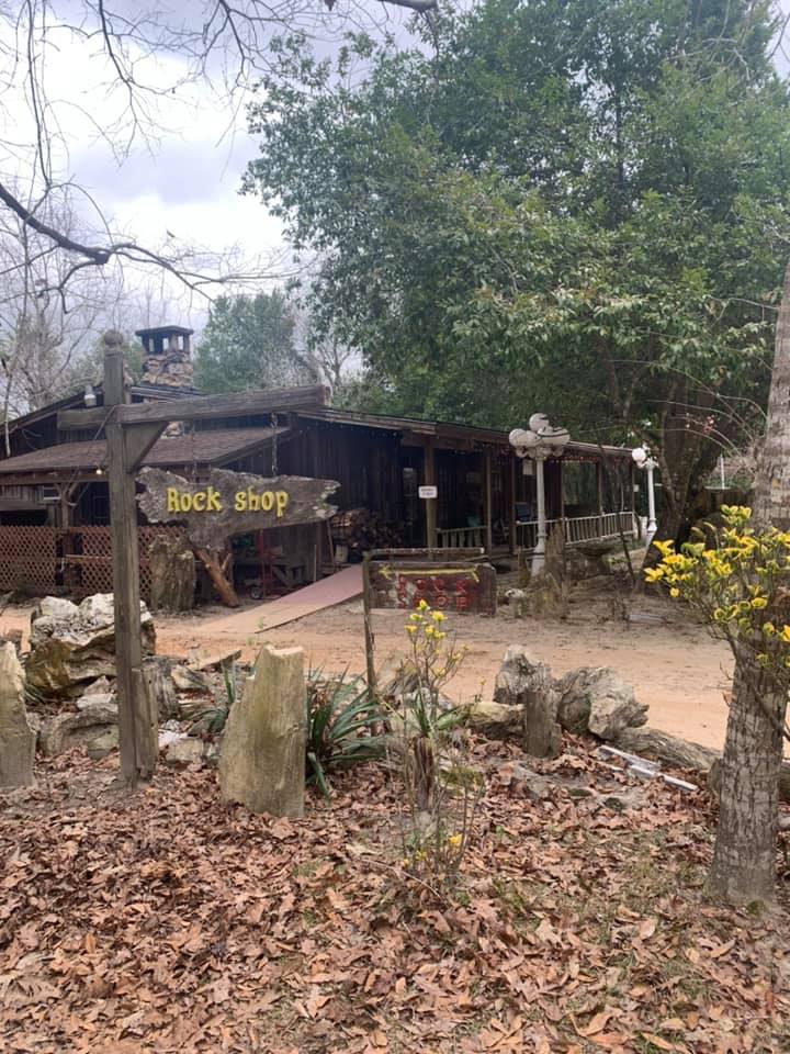 Rustic wooden shop in a wooded area with a “Rock Shop” sign, rocks, and leaf-covered ground.