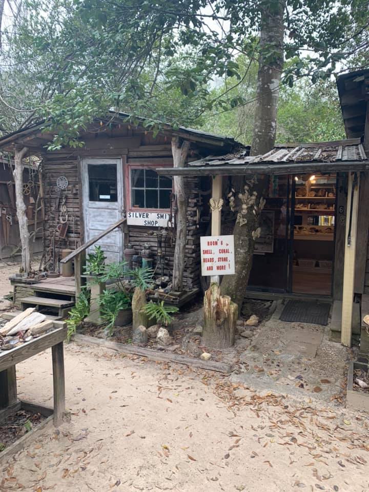 Small rustic roadside shop with wooden porch, hanging sign, and shelves inside among trees.