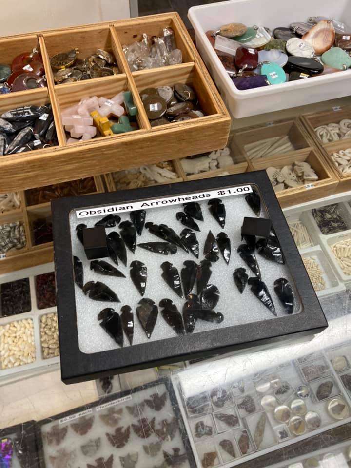 Trays of assorted gemstones and dark mineral specimens displayed in shallow wooden and plastic bins.