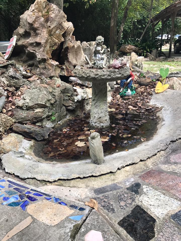 Small outdoor rock fountain with a shallow circular basin and colorful decorative figures beside a walkway.