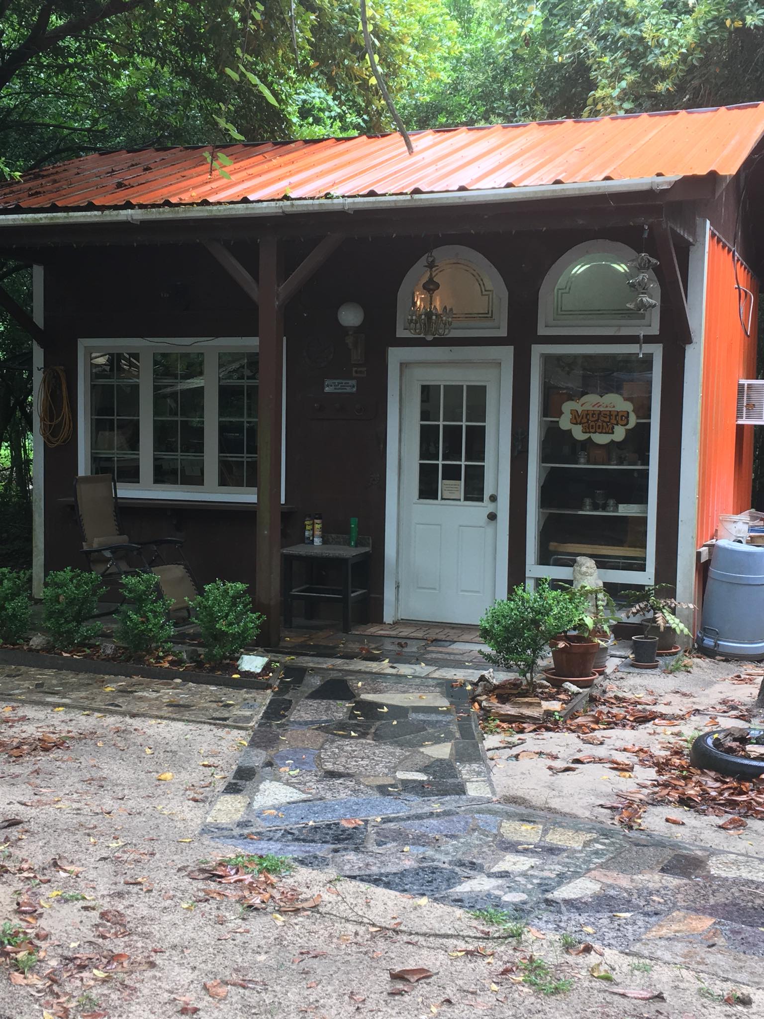 Small rustic cabin with a red metal roof, white door, and windows surrounded by trees and greenery