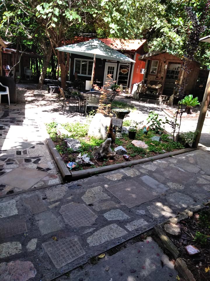 Stone path leading to a small rustic cabin amid trees and a flower bed in a shaded garden