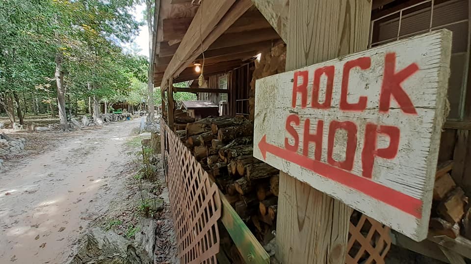 Rock shop sign beside a wooden roadside stall with stacked stones and a dirt path