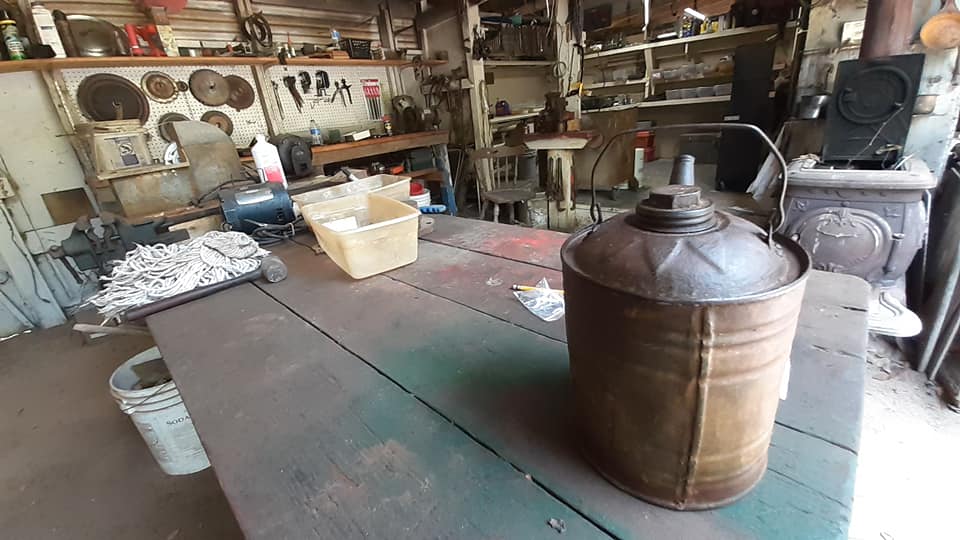Rustic workshop with a long wooden table, copper kettle in front, and shelves of tools and cookware.