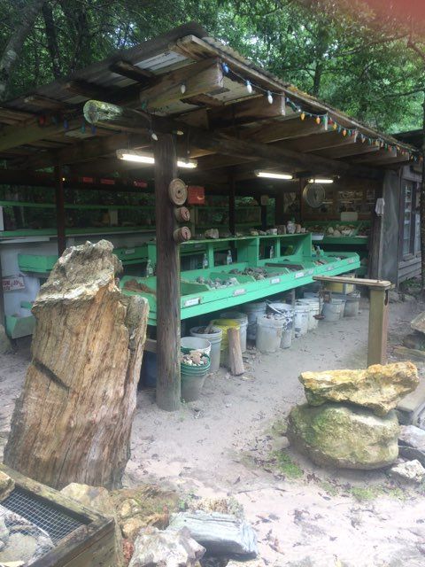 Outdoor rustic shelter with green workbenches and tools, surrounded by trees and rocks.