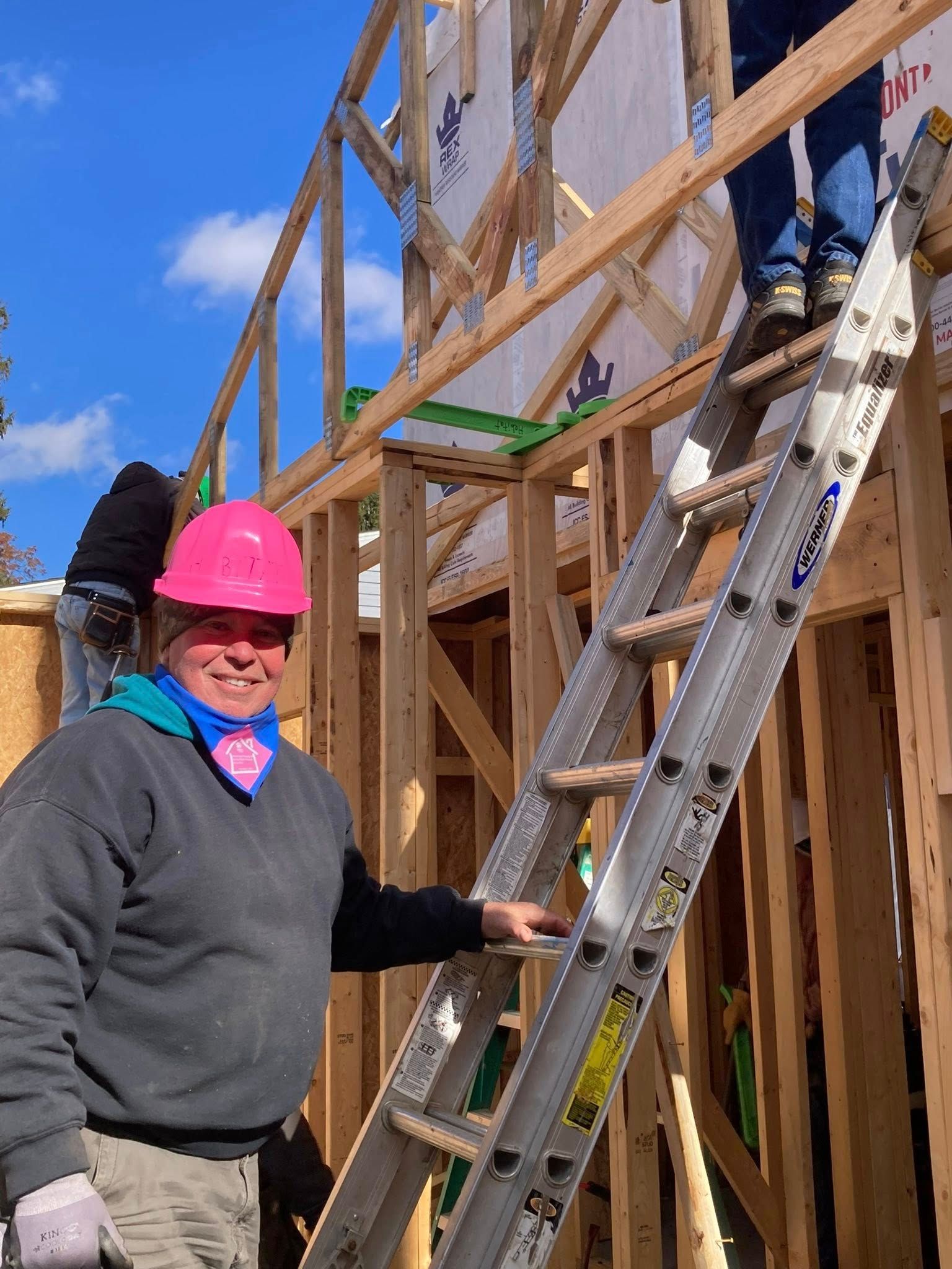 Worker in a pink hard hat standing by a ladder on a wooden frame construction site under a blue sky