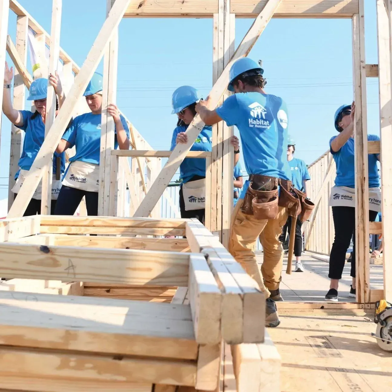 Construction crew in blue shirts and hard hats framing a wooden house in bright sunlight