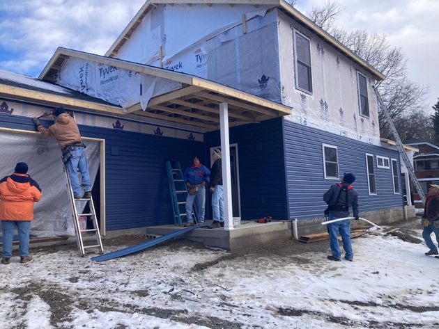 Workers repairing a blue house in winter, one man on a ladder under the porch roof.
