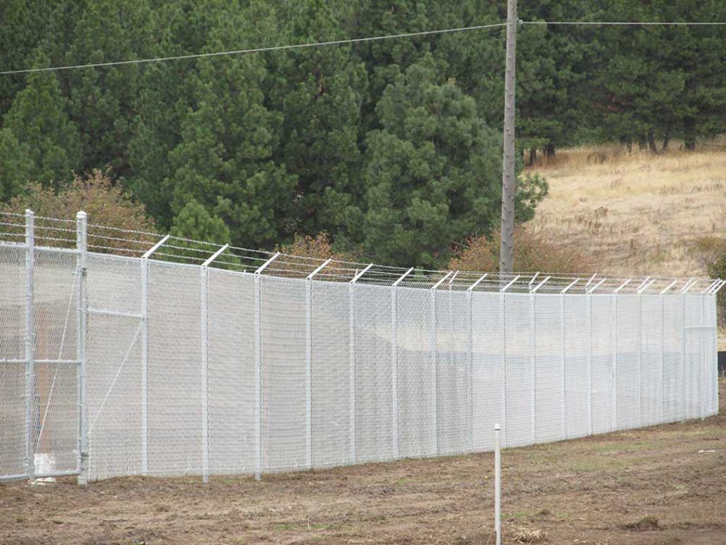 A fence with barbed wire surrounding a field with trees in the background.