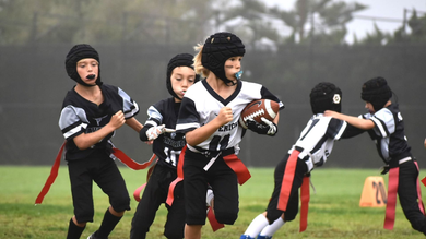 Flag football game at night, a player in red jersey runs with the ball as others pursue on a green field.