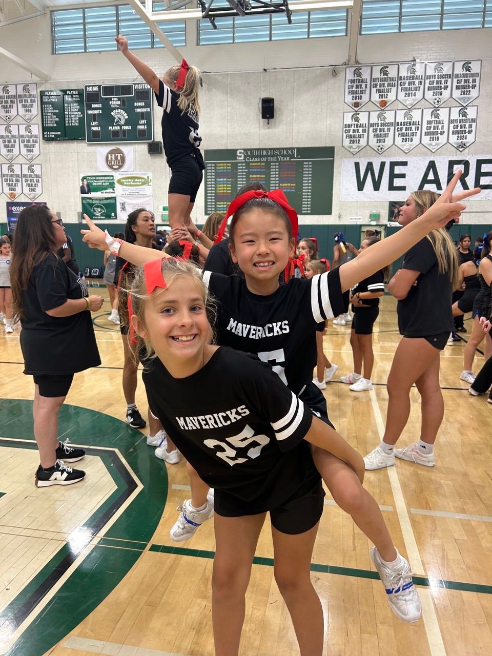 Youth volleyball team posing mid-routine in a gym, wearing black uniforms with red bows and arms outstretched.