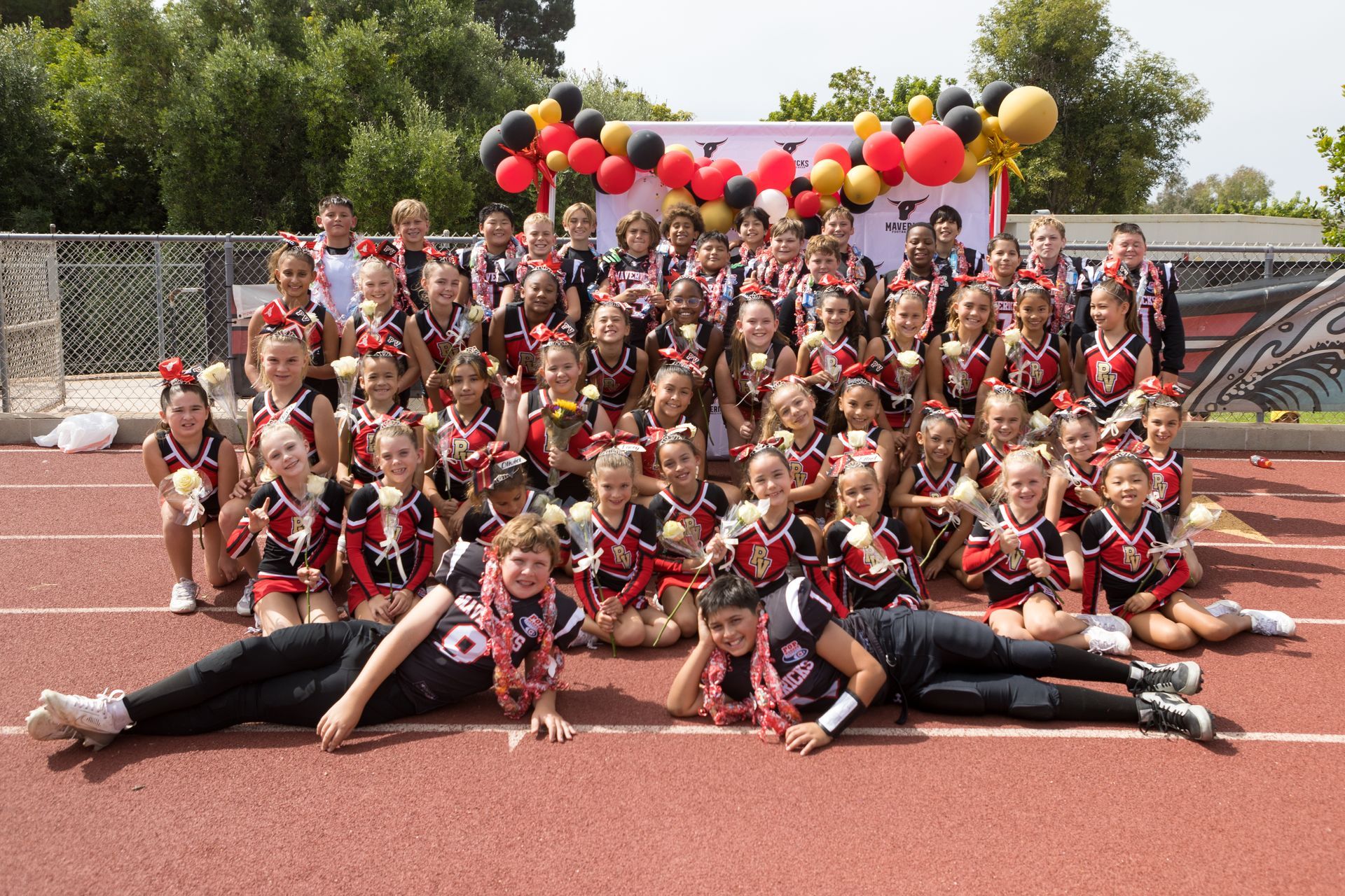 Large group posing on a track in red-and-black uniforms, with balloons overhead and two people lying in front