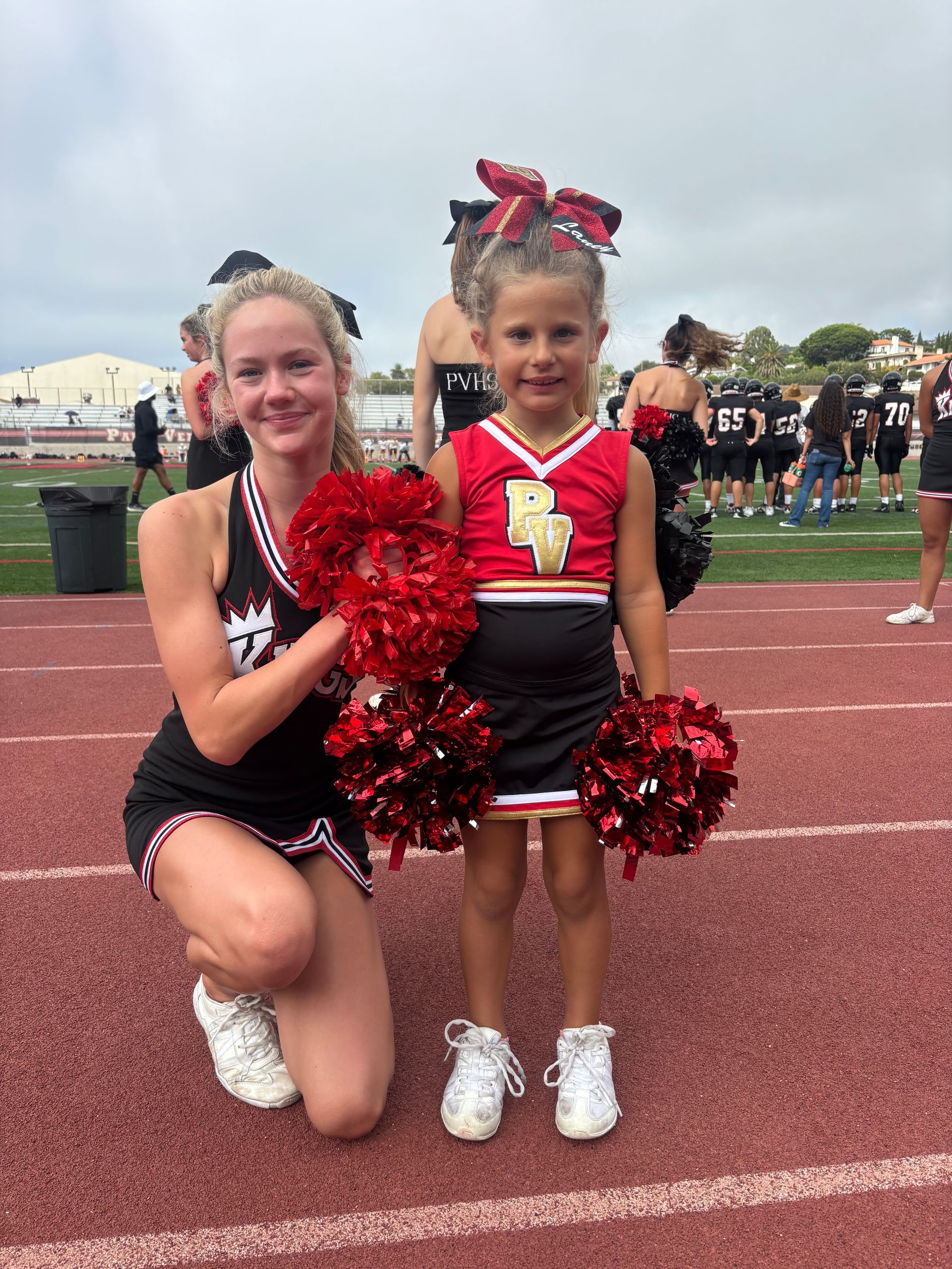Cheering girl and child in red-and-black uniforms kneel on a track holding red pom-poms at a sports event