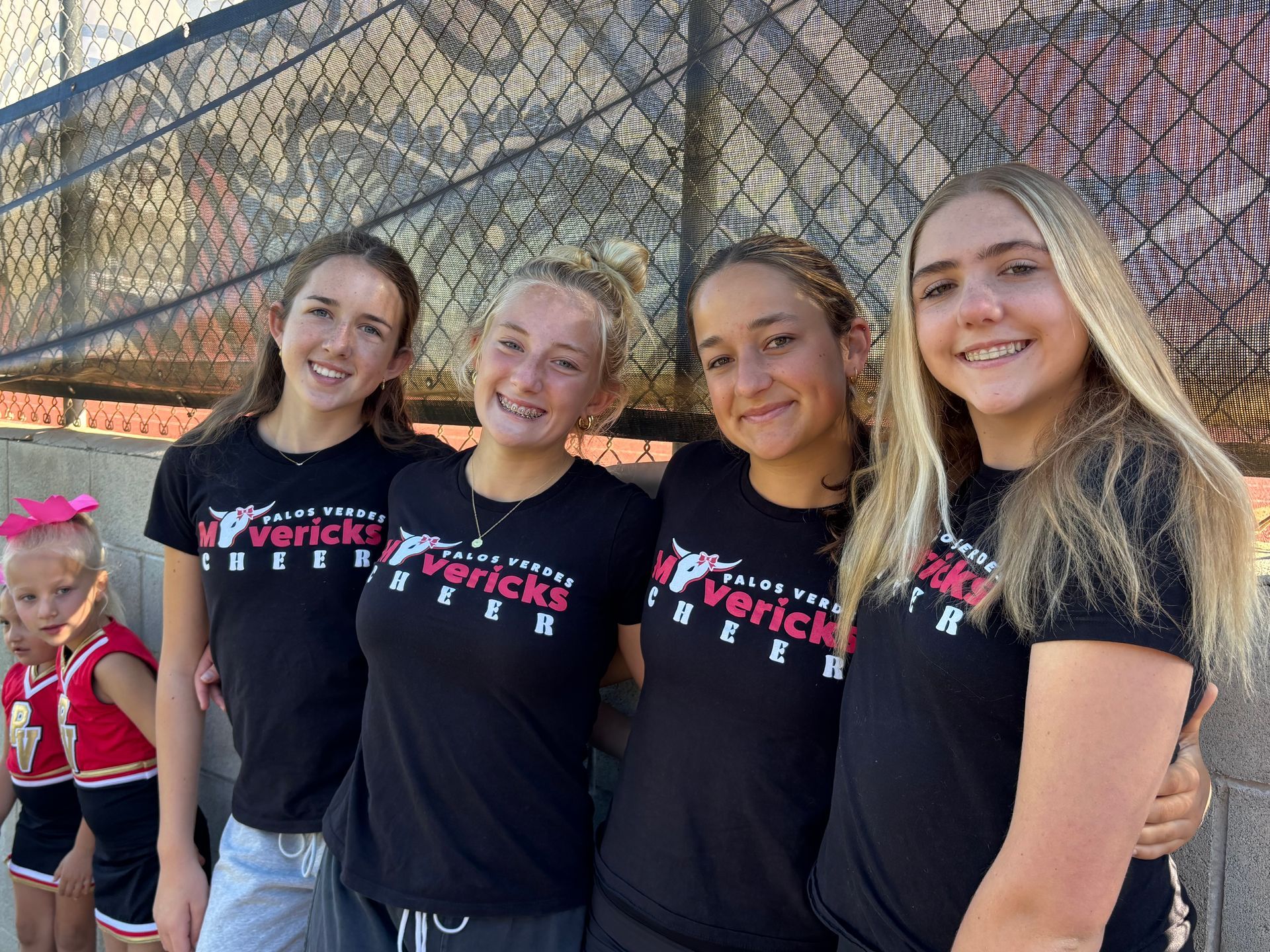 Four cheerleaders in black shirts posing and smiling in front of a chain-link fence outdoors