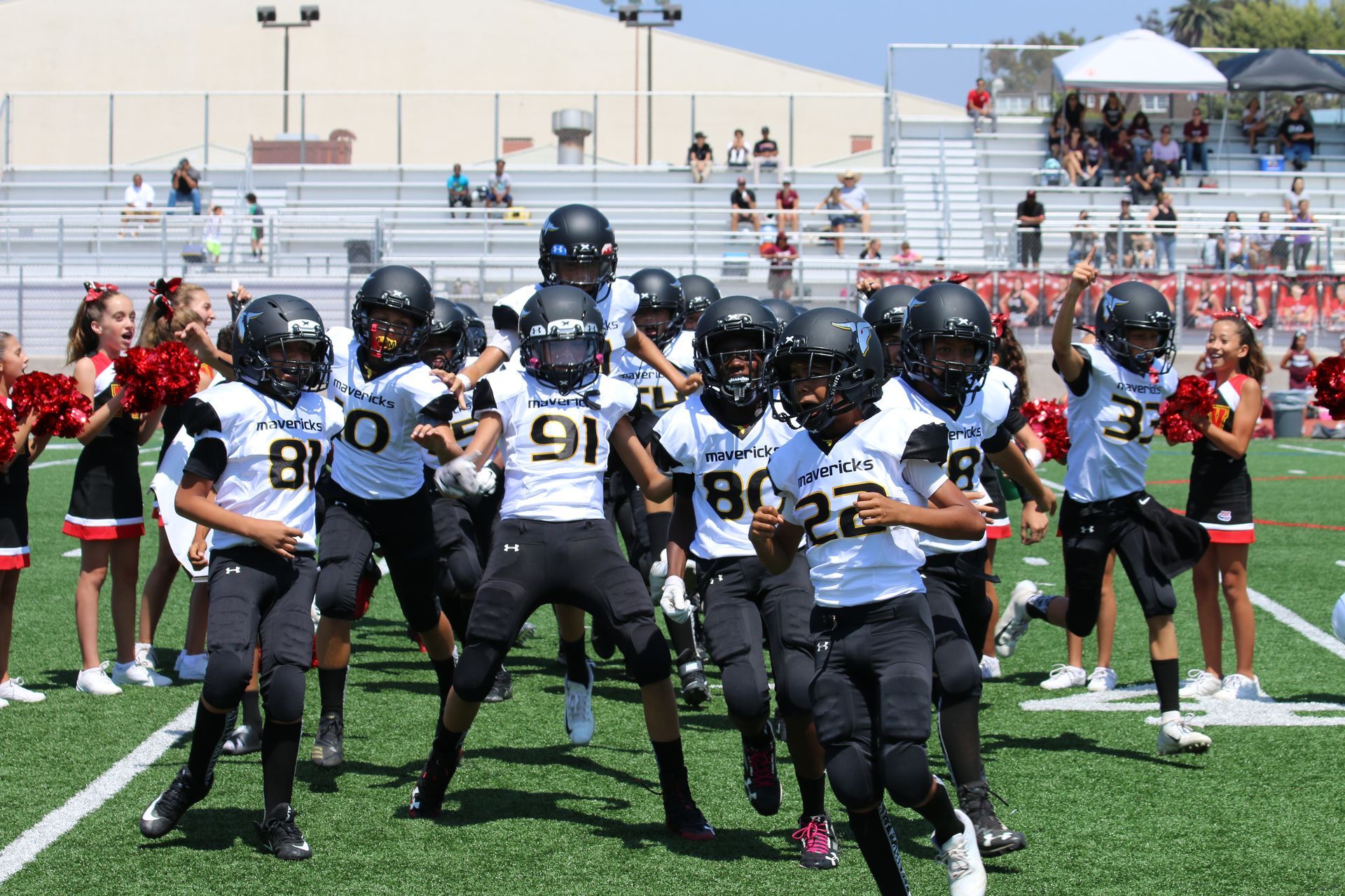 Youth football players in black and white uniforms huddle on a field during a game, with cheerleaders behind them.