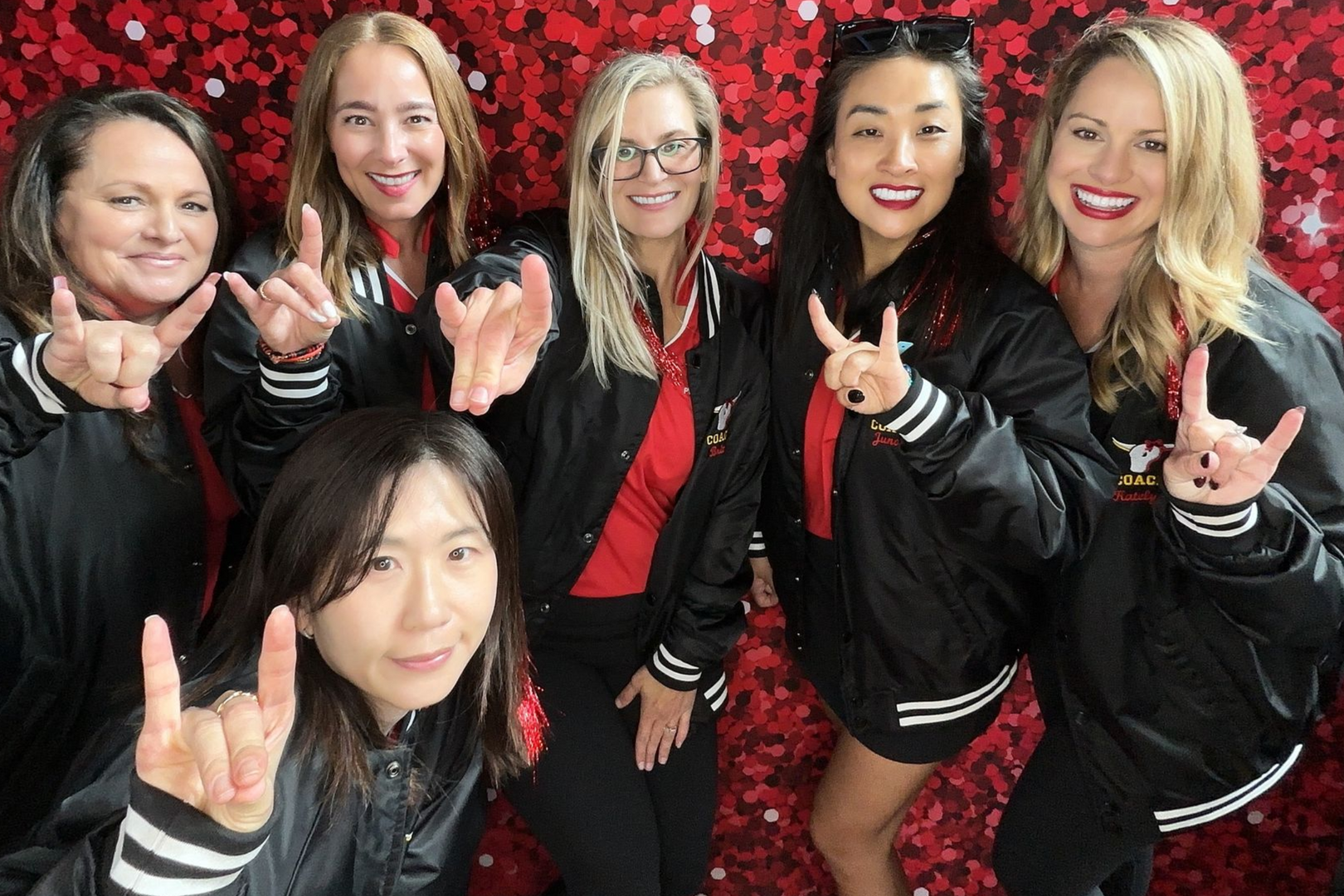 Six people posing together in black jackets against a red floral backdrop, smiling and making hand signs