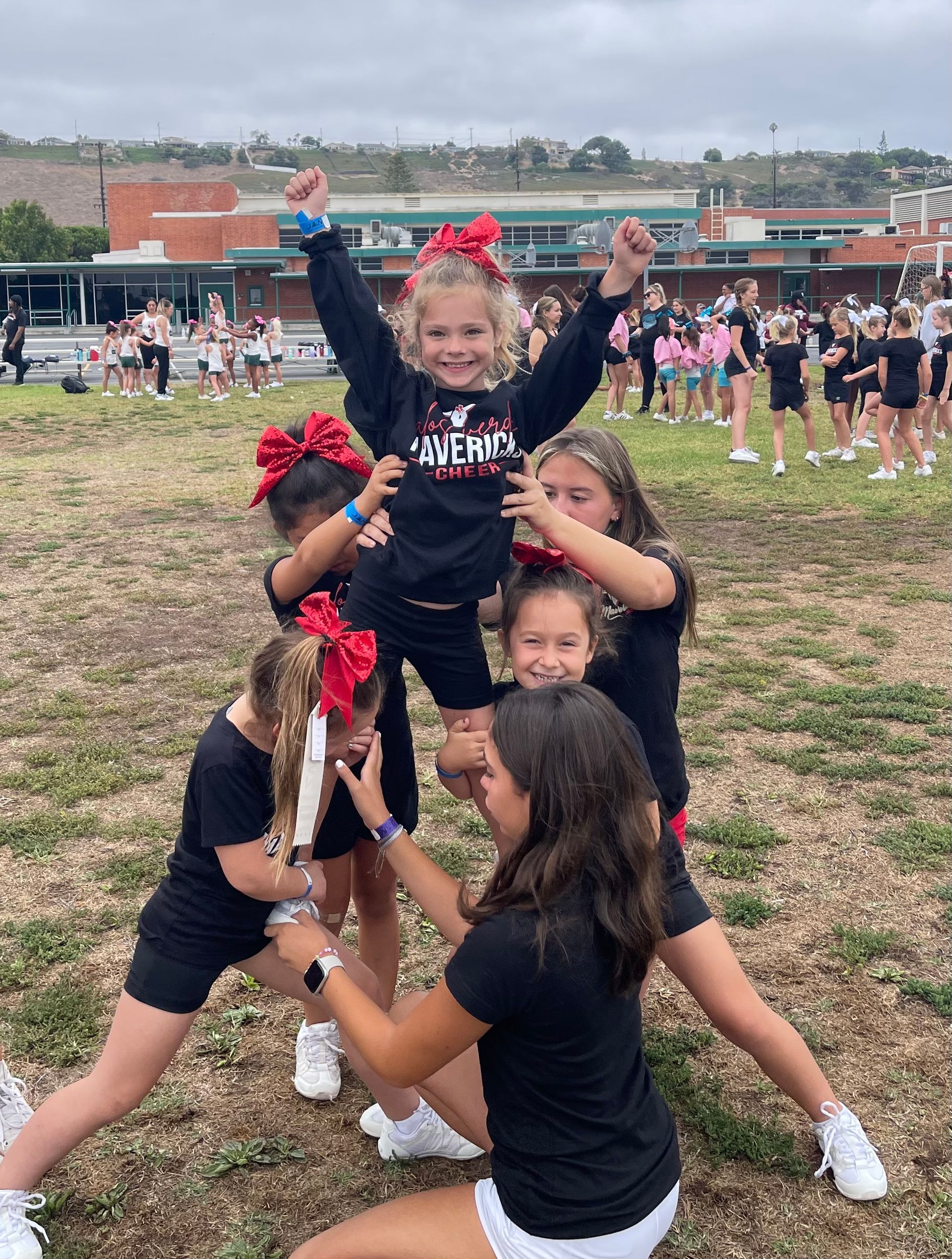 Cheerleaders in black uniforms pose in a pyramid on a grassy field at an outdoor event