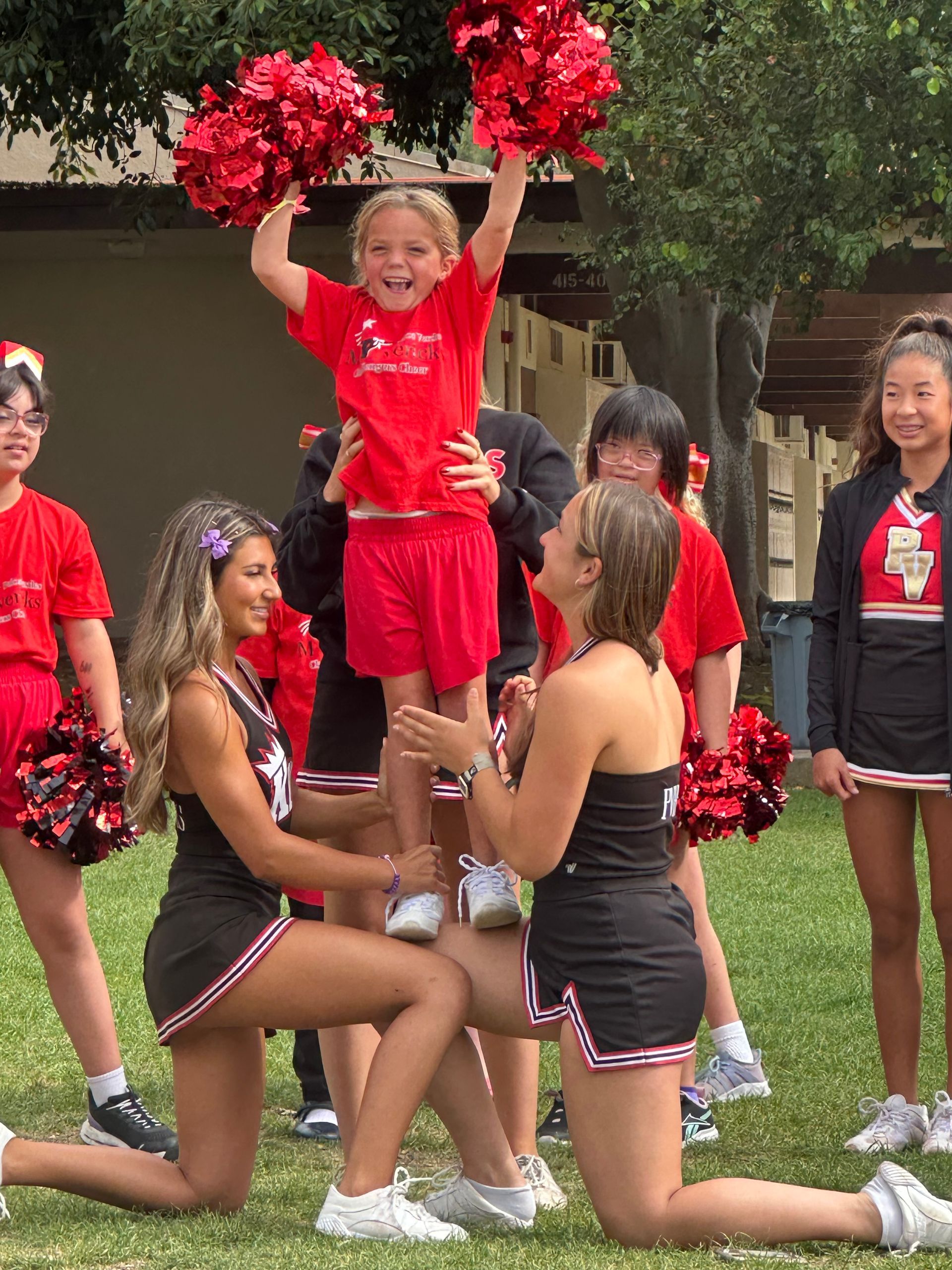 Cheerleaders in red and black uniforms lift a child holding red pom-poms on a grassy field.