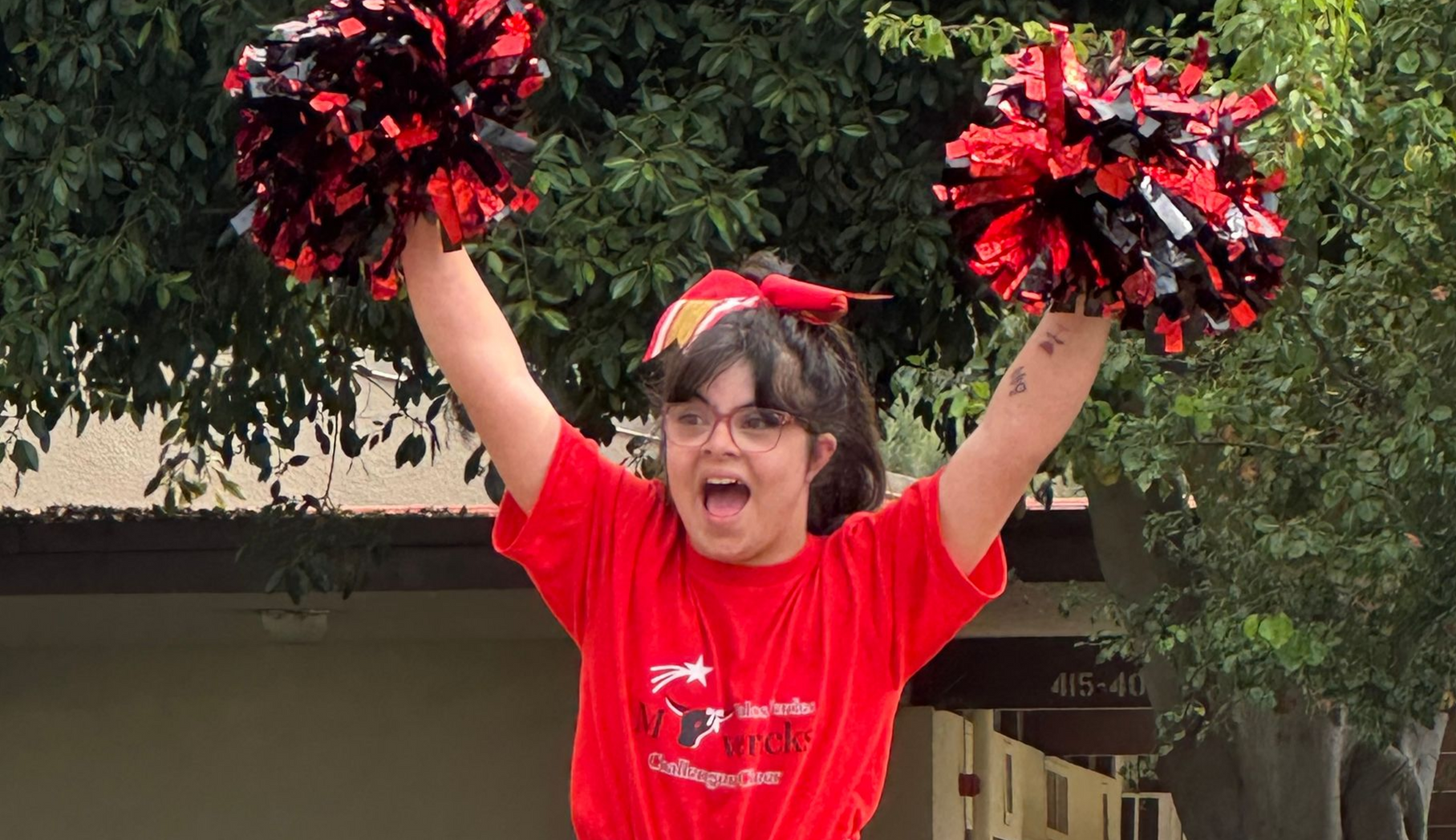 Person in a red shirt cheering with raised pom-poms outdoors under a tree