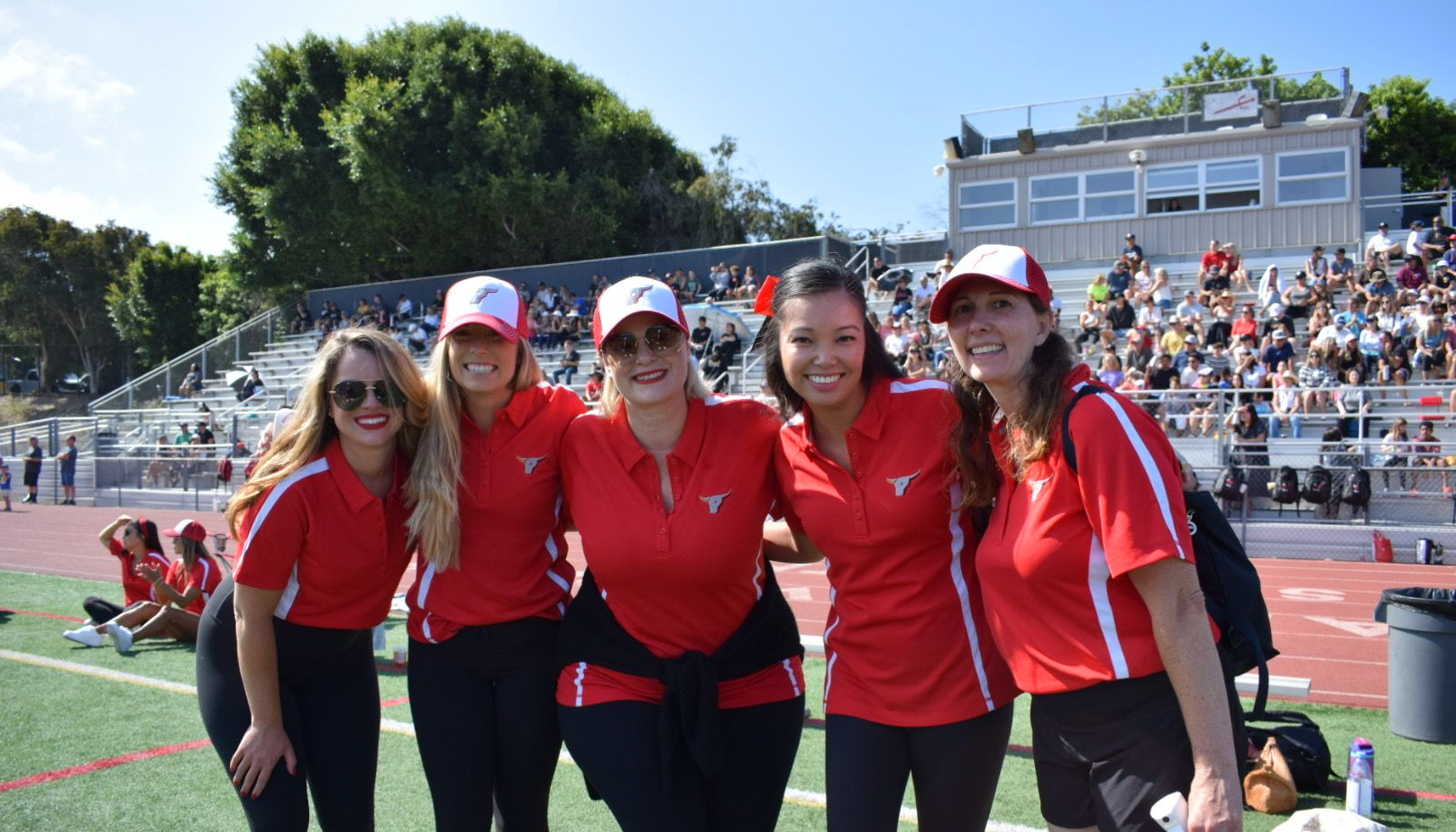 Five women in red shirts and white caps smiling on a sports field with a crowd in the stands