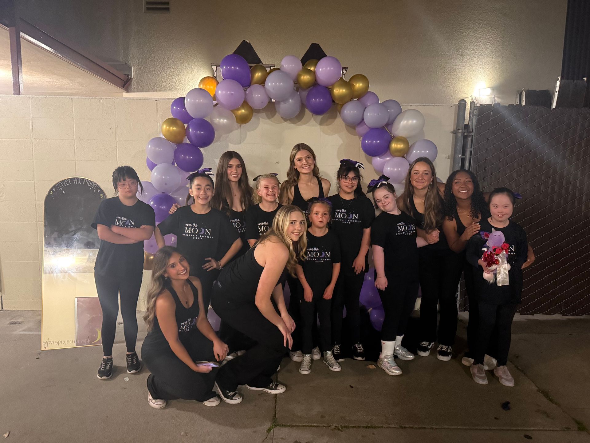 Group posing under purple and gold balloon arch at an outdoor celebration at night