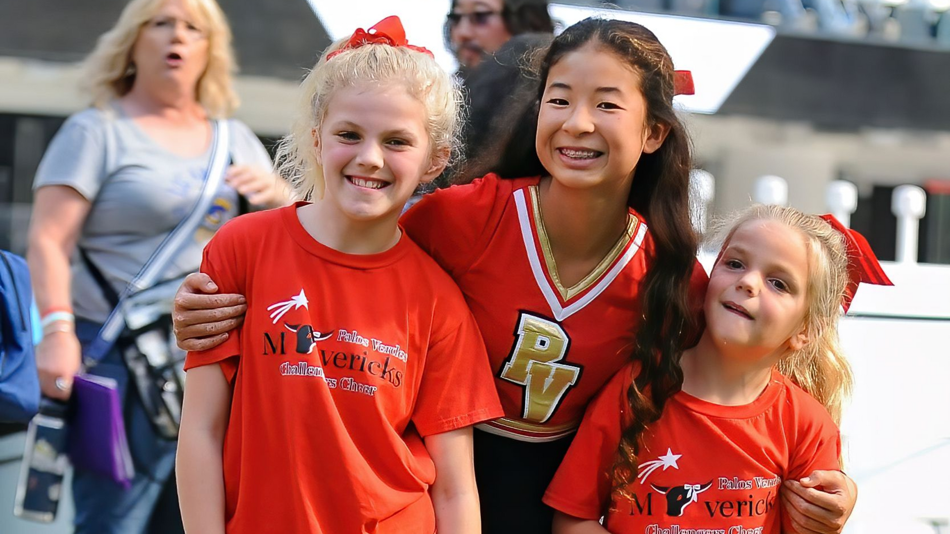 Three smiling cheerleaders in red team shirts pose outdoors, one in a black-and-red uniform with a letter P.