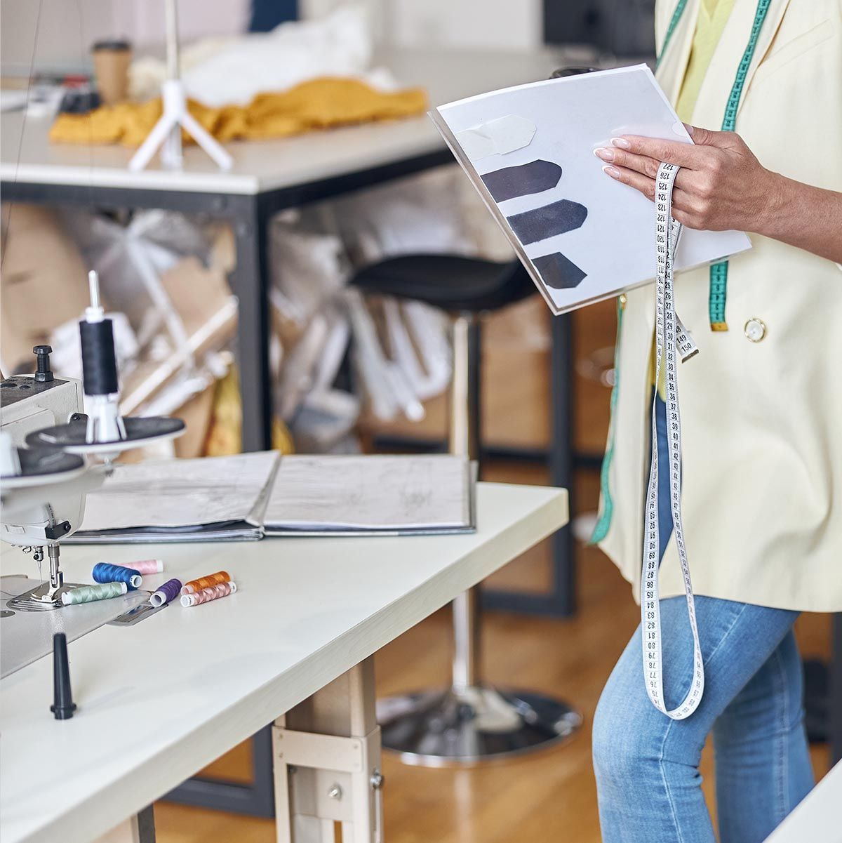 Person holding a notebook and measuring tape in a sewing studio, with a sewing machine in the foreground.