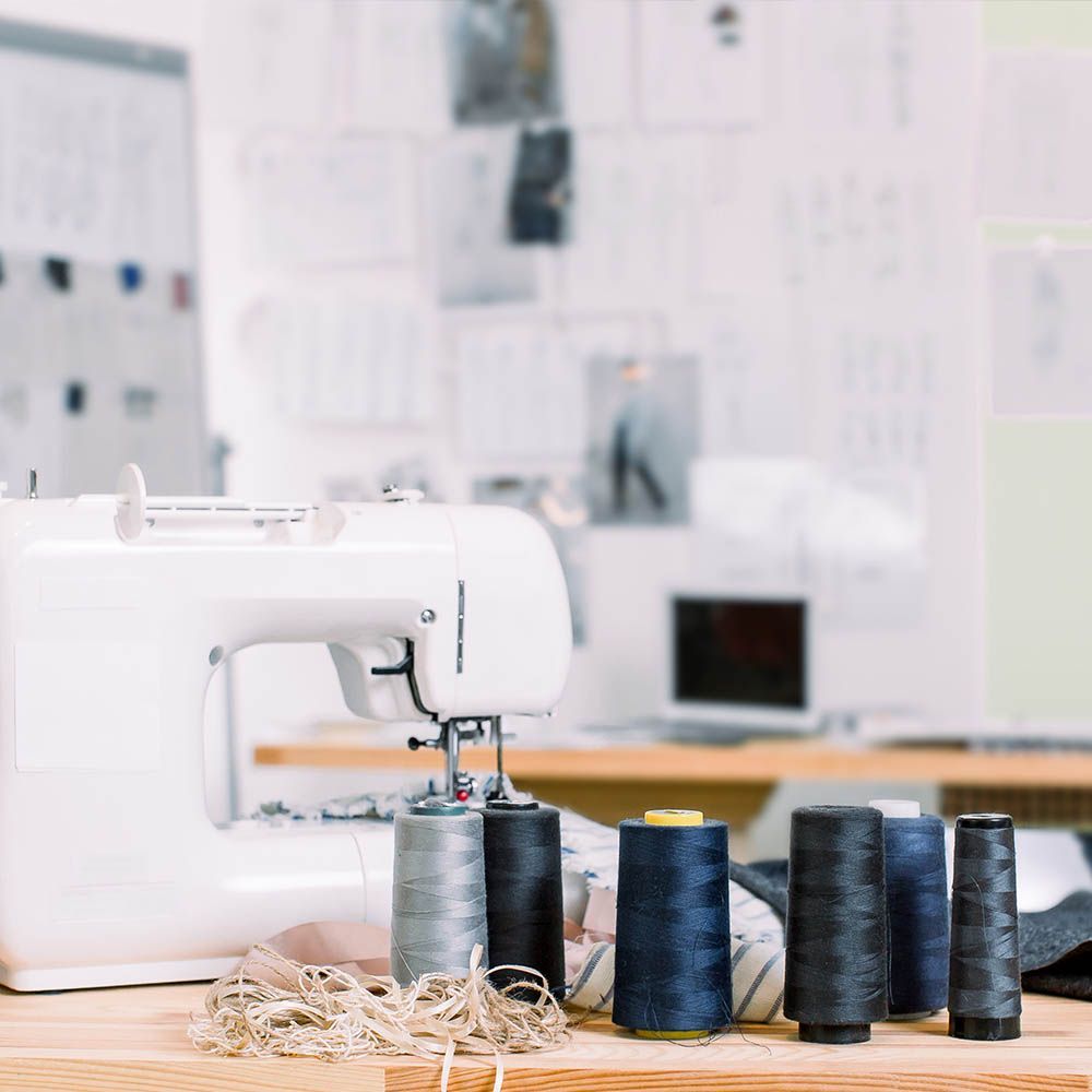 Sewing machine and spools of thread on a wooden table, with sketches and a laptop in the background.