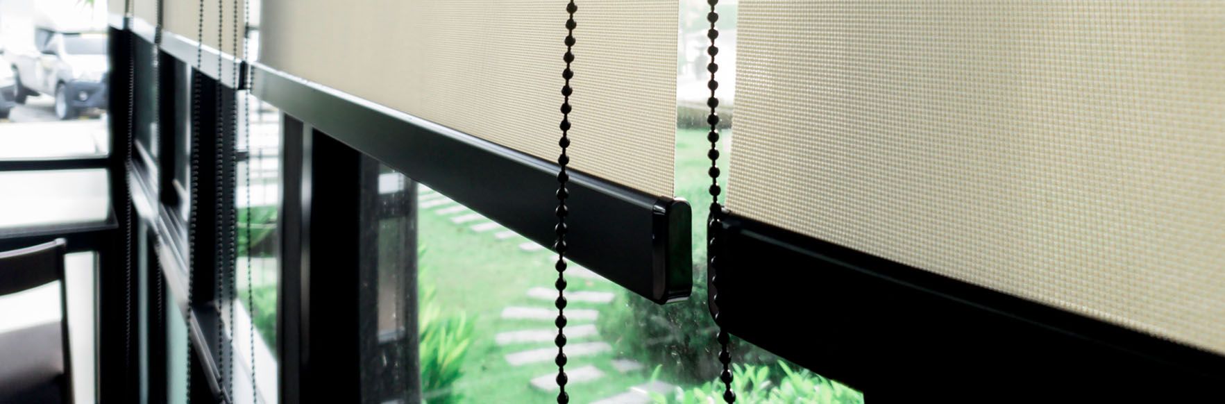 Close-up of beige roller blinds with black hardware and beaded pulls, covering a window overlooking greenery.