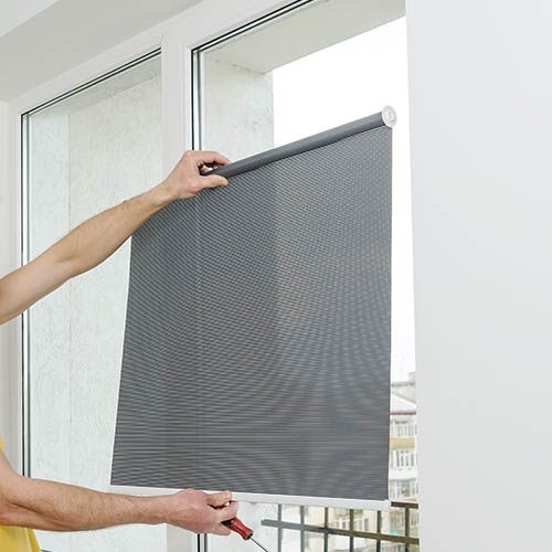 Person installing a gray roller shade on a white window frame, outdoors visible.